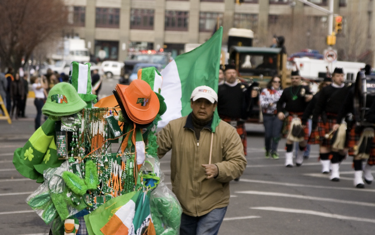 A street vendor selling festive St. Patrick’s Day accessories in a cart, down the Hoboken St. Patrick's Day parade, as men wearing traditional Irish kilts, holding bagpipes, walk in the parade in unison. Photo courtesy of Flickr
