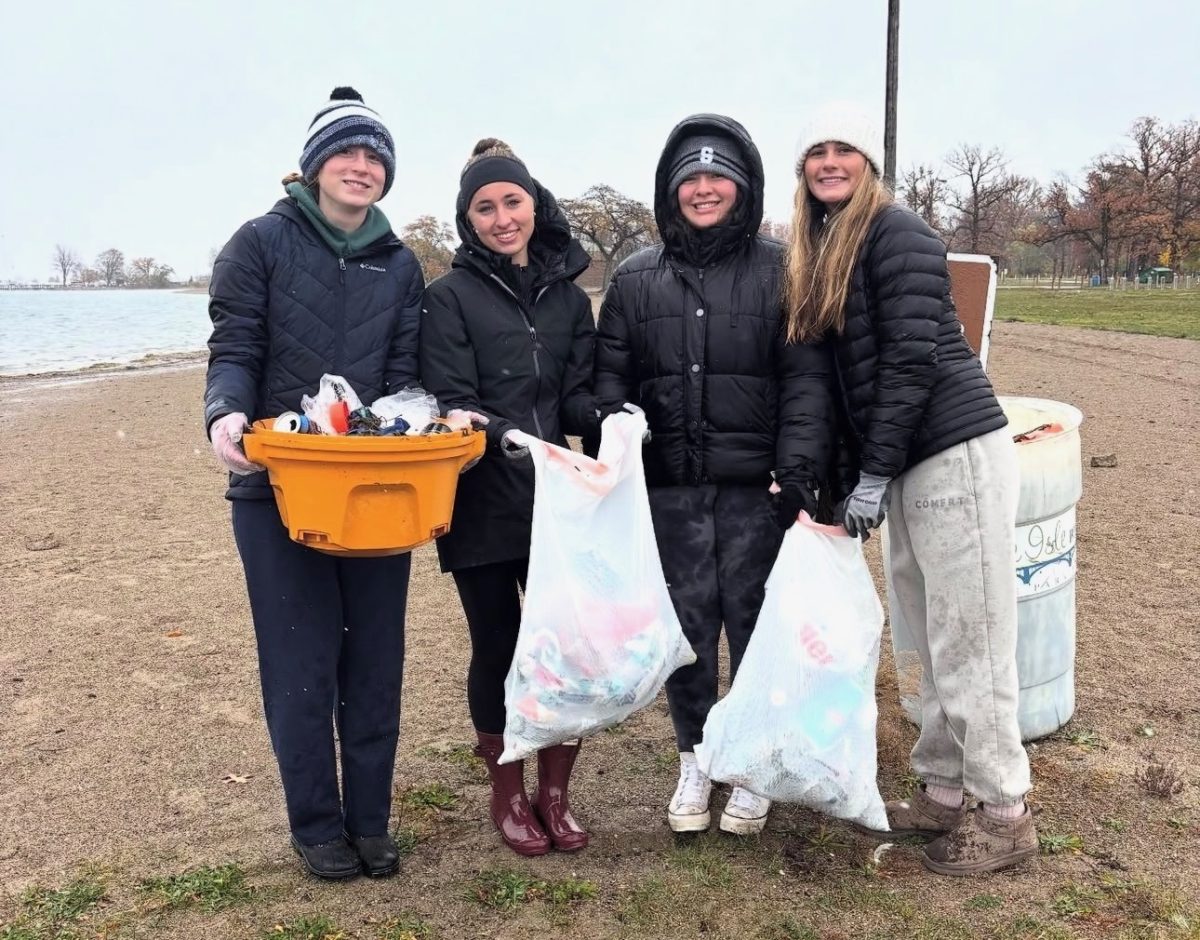 Save the Lakes cleans up Belle Isle beaches for a community conservation project. Chloe Slawson ’26 (left), Abby Macey ’26, Mackenzie Adams ’26 and Morgan Mannino ’26 (right) hold up trash bags filled with garbage left behind by beach goers.